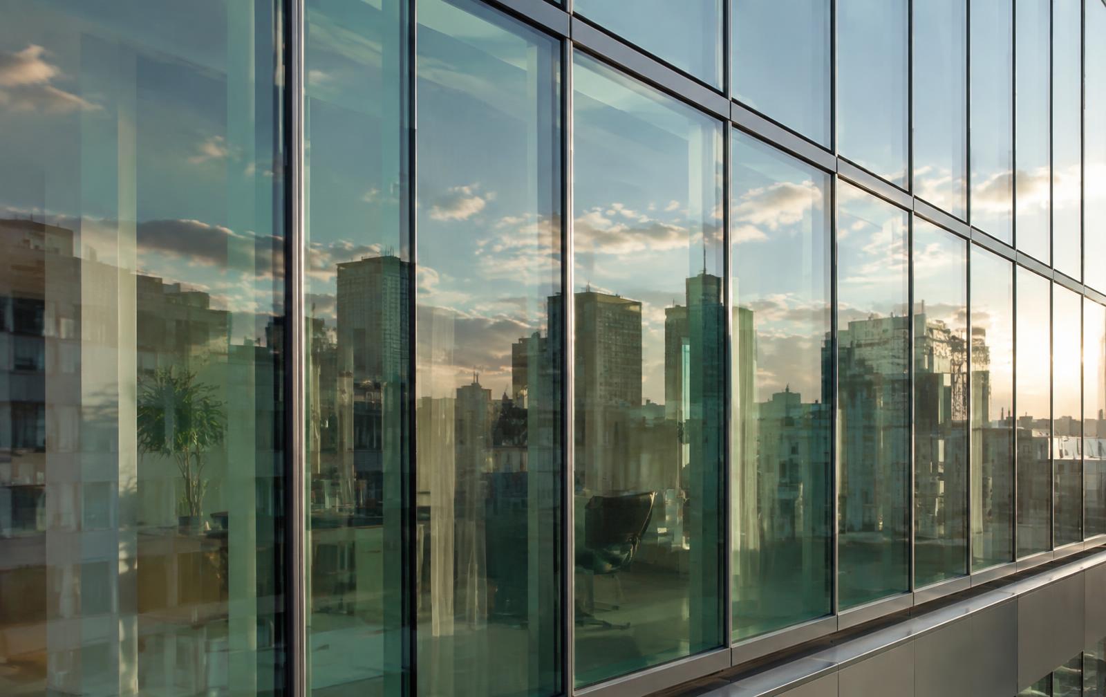 Close up render of a glazed office facade in late afternoon light, showing green tinted double glazing, reflections of neighbouring buildings and sky, and faint interior detail behind the glass